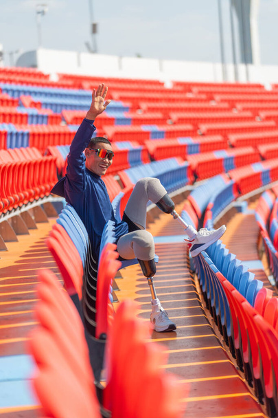 Image verticale vue de côté de sport homme athlète prothèse jambes s'asseoir sur la chaise de l'amphithéâtre regarder vers l'avant avec spectacle action de bonheur en levant les deux mains vers le haut dans le stade avec la lumière du jour. - Photo, image