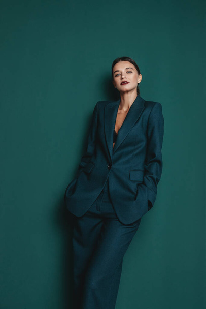 Fashionable confident woman wearing elegant green suit with blazer and trousers posing on green wall background. Studio fashion portrait - Foto, afbeelding
