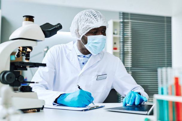 Porträt eines erwachsenen schwarzen Mannes als Wissenschaftler, der Laptop benutzt, während er im medizinischen Labor forscht - Foto, Bild