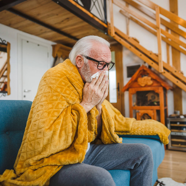 ένας καυκάσιος ηλικιωμένος άνδρας κάθεται στον καναπέ-κρεβάτι στο σπίτι κρατώντας χαρτομάντιλο κοινό κρύο φτάρνισμα - Φωτογραφία, εικόνα