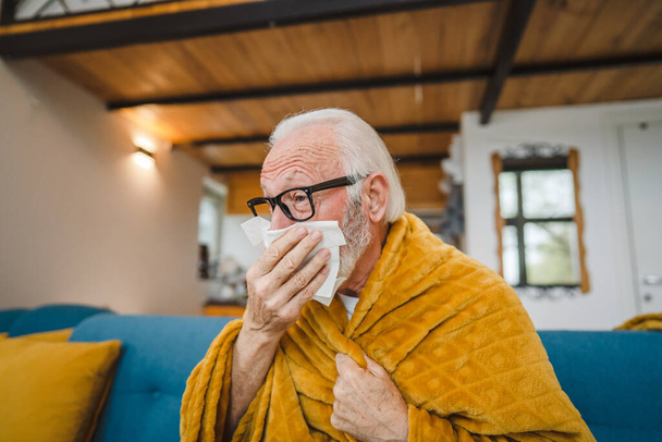 un homme âgé caucasien assis sur le canapé-lit à la maison tenir papier tissu commun froid éternuer - Photo, image