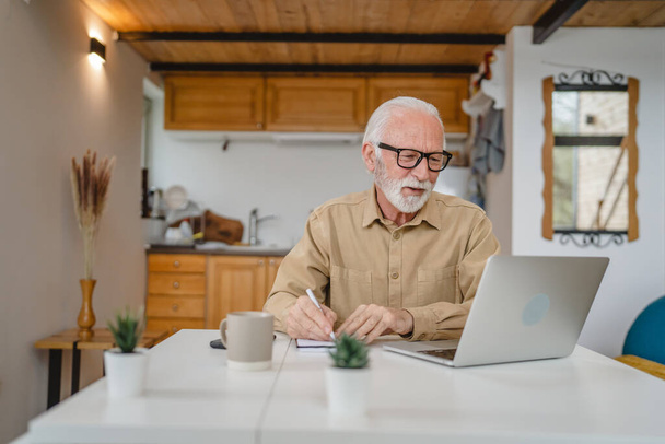 Ένας ηλικιωμένος καυκάσιος παππούς με γενειάδα και γυαλιά εργάζονται σε φορητό υπολογιστή χρήση στο σπίτι σε απευθείας σύνδεση ευτυχής χαμόγελο επιτυχία - Φωτογραφία, εικόνα