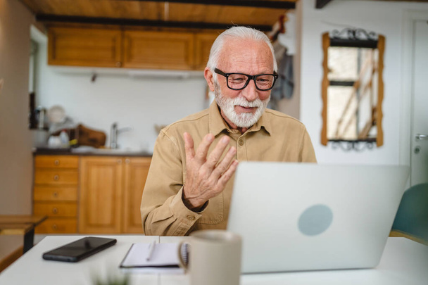 Ανώτερος καυκάσιος άντρας χρησιμοποιεί φορητό υπολογιστή για να κάνει μια βιντεοκλήση στο σπίτι - Φωτογραφία, εικόνα
