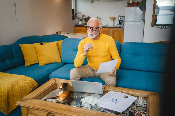 Ein älterer kaukasischer Großvater mit Bart und Brille arbeitet zu Hause am Laptop - Foto, Bild