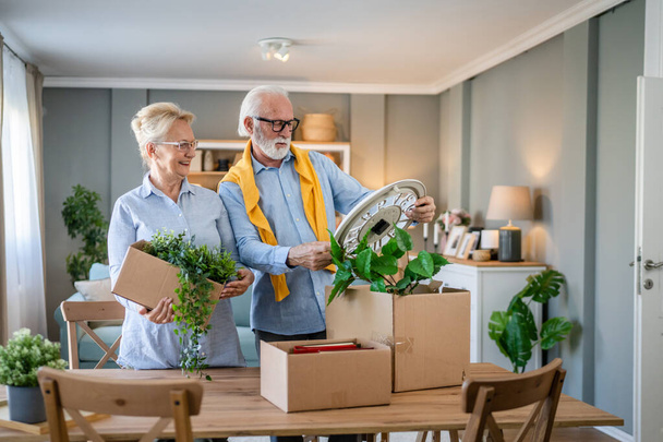 family senior couple man woman husband and wife pensioner grandfather grandmother moving in new apartment taking their stuff belongings and plants in or out of boxes packing or unpacking real people - Fotoğraf, Görsel