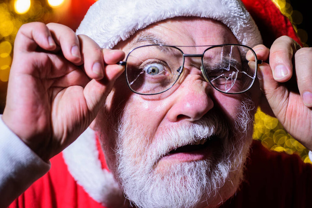 Vánoce. Santa Claus s rozbitými brýlemi. Vousatý muž v kostýmu Santy se zlomenými pánskými brýlemi. Santa se dívá popraskanými brýlemi. Překvapený Santa Claus s lámanými brýlemi - Fotografie, Obrázek