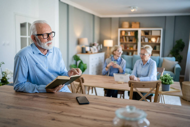 Un hombre adulto mayor caucásico pensionista masculino moderno abuelo con pelo corto y barba leer libro en casa usar gafas de vista persona real copiar espacio - Foto, Imagen