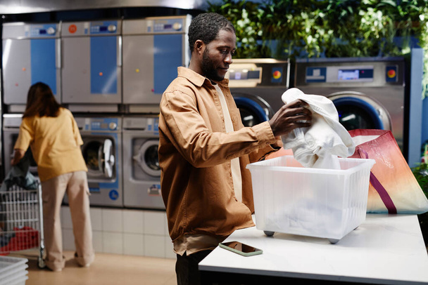 Afroamerykanin w stroju casualwear przygotowuje brudne ubrania do prania maszynowego stojąc przy stole i wkładając je do pojemnika - Zdjęcie, obraz