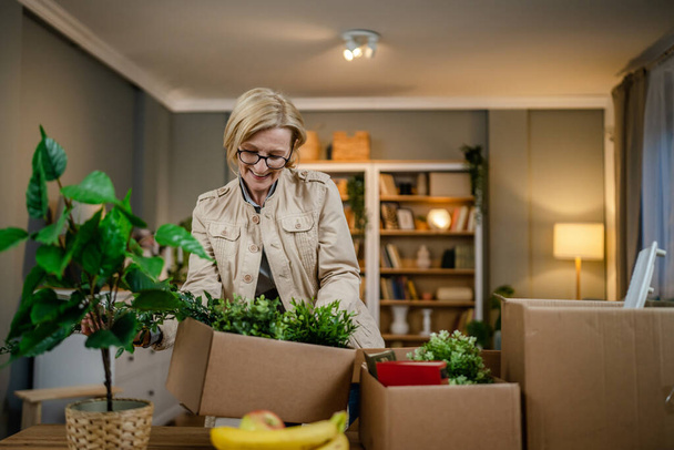 een oudere vrouw gepensioneerde oma verhuizen in een nieuw appartement nemen haar spullen en planten in of uit dozen inpakken of uitpakken echte persoon kopieerruimte - Foto, afbeelding
