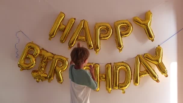 Счастливый мальчик 6-8 смотрит на надпись на стене Счастливого дня рождения из золотых шариков, прикасается руками к письмам. Готовлюсь к празднованию дня рождения. Любовь и забота. Предвкушение праздника. 4k кадров - Кадры, видео