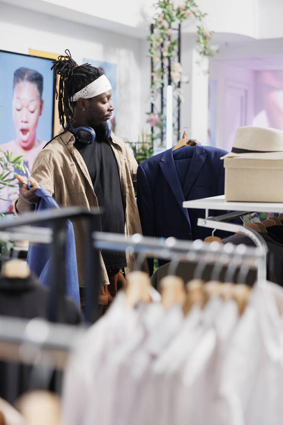 Afroamerikaner mit zwei Jacken auf Kleiderbügeln, die Stil und Passform prüfen. Käufer wählt formalen Blazer, während er in der Nähe von Regalen voller Kleidung steht und Kleidung im Einkaufszentrum einkauft - Foto, Bild