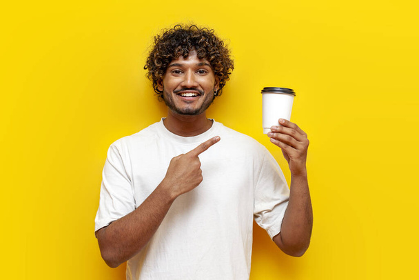 mladý indián ukazuje na papírový šálek kávy na žlutém izolovaném pozadí, jihoasijský muž v bílém tričku ukazuje a reklamní nápoj - Fotografie, Obrázek