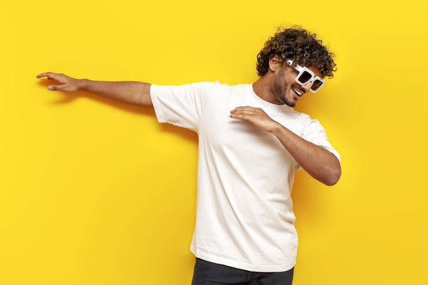 young indian guy in sunglasses dancing dab on yellow isolated background, south asian man in white t-shirt raises his hands up and shows dab gesture - Photo, Image