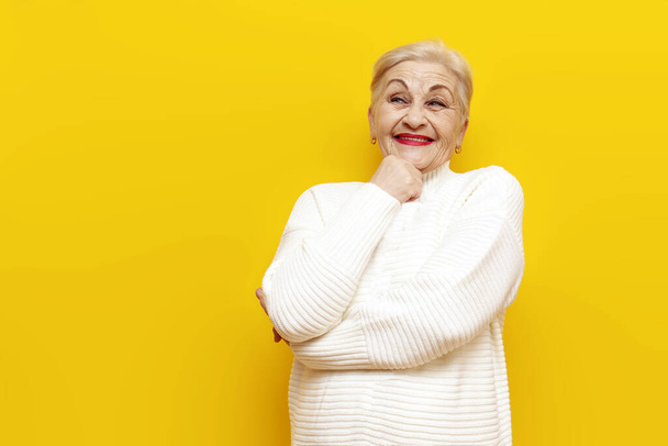 Beyaz kazaklı düşünceli yaşlı bir büyükanne sarı izole bir arka planda hayal eder ve gülümser. Kırışıklıkları olan yaşlı bir kadın bir fikre bakıp düşünür ve güler. - Fotoğraf, Görsel