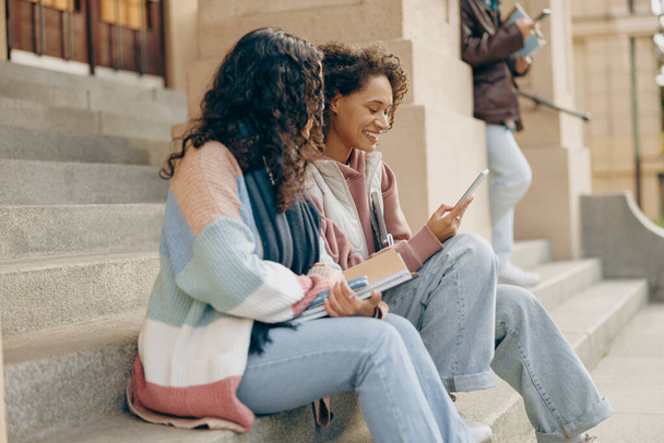 Группа молодых счастливых студентов смотрит что-то за мобильным телефоном, сидящим на лестнице - Фото, изображение