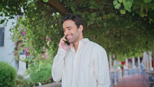 Um tipo a rir a ligar para o telemóvel no aconchegante beco da natureza. Bonito homem sexy falando smartphone esperando namorada na data da cidade. Unshaven feliz macho bate-papo celular sorrindo sob árvore verde - Filmagem, Vídeo