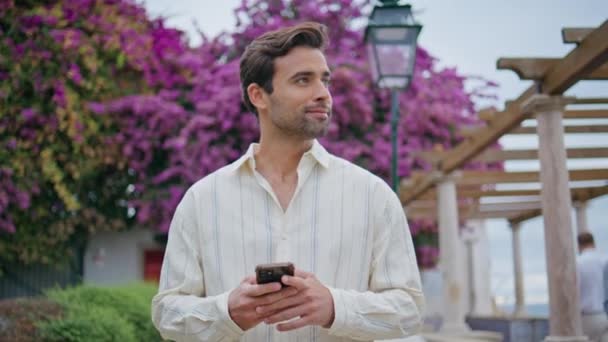 Sexy macho drží mobilní telefon v květu stromů uličky detailní. Mladý neoholený muž skrývající smartphone procházející se v letním parku. Hezká italská brunetka procházky sám relaxační na bulváru zeleně  - Záběry, video