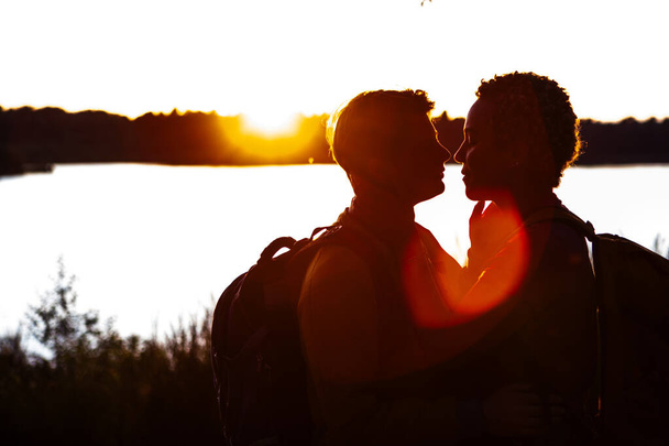 Dieses eindrucksvolle Bild zeigt die Silhouetten zweier Menschen, die einen intimen Moment vor dem Hintergrund einer untergehenden Sonne über einem ruhigen See teilen. Ihre Profile werden durch die warmen Farbtöne der - Foto, Bild