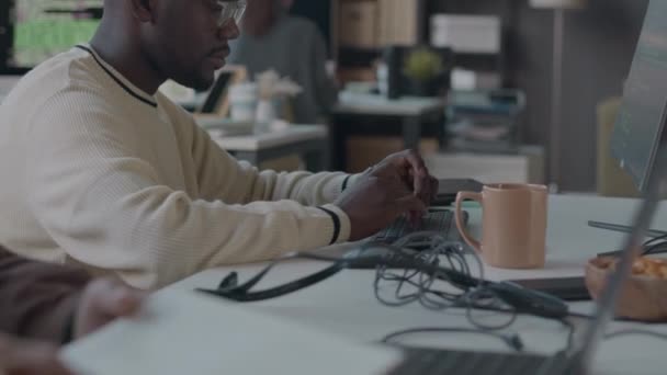Cintura para cima do jovem desenvolvedor de software masculino preto digitando no teclado sem fio enquanto trabalhava em escritório de programadores modernos com colegas - Filmagem, Vídeo