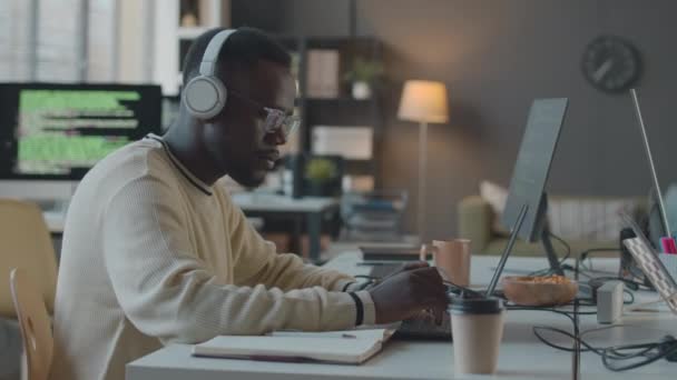 Талия с видом на боковую сторону снимок молодого афроамериканского программиста, одетого в беспроводные наушники и очки, работающего на ноутбуке в минималистском офисе - Кадры, видео