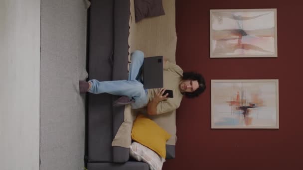 Vertikale Langzeitaufnahme eines jungen kaukasischen Freiberuflers mit Laptop und Anrufbeantworter auf dem Smartphone, während er auf der Couch im stilvollen Wohnzimmer sitzt und von zu Hause aus arbeitet - Filmmaterial, Video