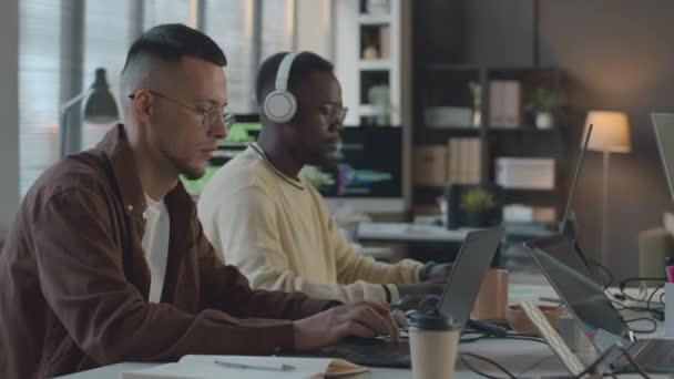Derékig két fiatal multiracionális szoftver mérnök dolgozik laptop és számítógép, miközben ül egy asztalnál minimalista programozók iroda - Felvétel, videó