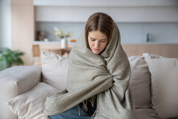 Depressive unglückliche Frau, die sich in kariertes Kleid vor Kälte hüllt, zittert, sich krank fühlt, krank ist, hohe Temperaturen hat. Frauen leiden unter Grippesymptomen, Entzündungen, saisonalen Viren, die Zeit zu Hause verbringen - Foto, Bild