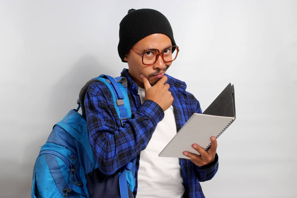 Estudiante asiático pensativo, con anteojos, sombrero de gorro y ropa casual, llevando mochila, se levanta sobre un fondo blanco, sosteniendo un libro y apoyando su mano sobre su barbilla en una pose reflexiva - Foto, imagen