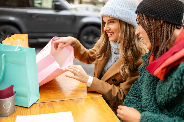 Duas mulheres alegres revelam seus achados de compras em uma mesa de café ao ar livre, desfrutando do prazer de novas compras. - Foto, Imagem