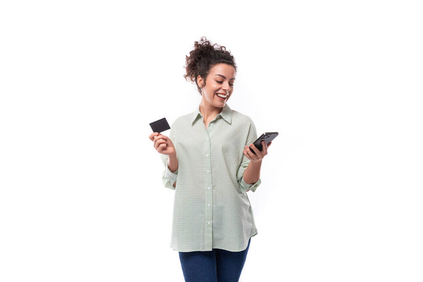 pretty young black-haired curly woman with casual bun styling in shirt holding plastic credit card mockup and smartphone. - Photo, Image