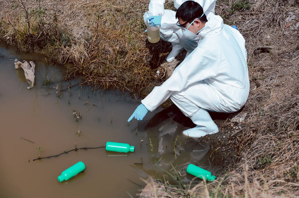 Zespół naukowców lub biologów nosi odzież ochronną do kontroli butelek z chemikaliami śmieci na naturalnym źródle wody i pobierania próbek wody. Koncepcja zanieczyszczenia wody. - Zdjęcie, obraz