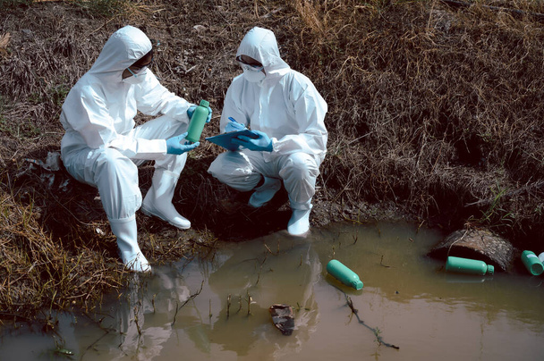 El equipo de científicos o biólogos usa ropa protectora para inspeccionar botellas de basura llenas de químicos en la fuente de agua natural y nota. Concepto de contaminación del agua. - Foto, imagen