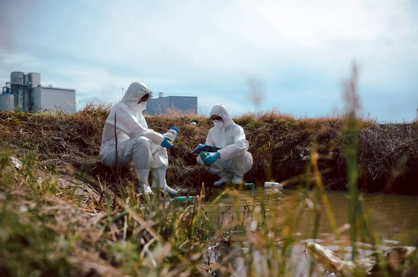 Ein Team von Wissenschaftlern oder Biologen trägt Schutzkleidung, um mit chemisch gefüllten Müllflaschen Wasserproben aus einer natürlichen Wasserquelle zu entnehmen. Wasserverschmutzungskonzept. - Foto, Bild