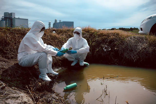 Team di scienziati o biologi indossa indumenti protettivi per ispezionare bottiglie piene di sostanze chimiche di rifiuti sulla fonte di acqua naturale e nota. Concetto di inquinamento idrico. - Foto, immagini