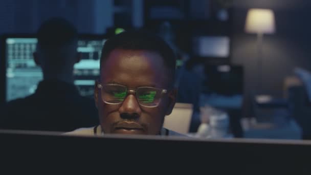 Fiatal fekete férfi kiberbiztonsági dolgozó dolgozik számítógép sötét irodában éjszaka zöld program kód fekete háttér tükröződik a szemüvegét - Felvétel, videó