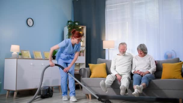örömteli női szociális munkás segít egy idős párnak, és porszívóval takarítja ki a padlót a lakásban. - Felvétel, videó