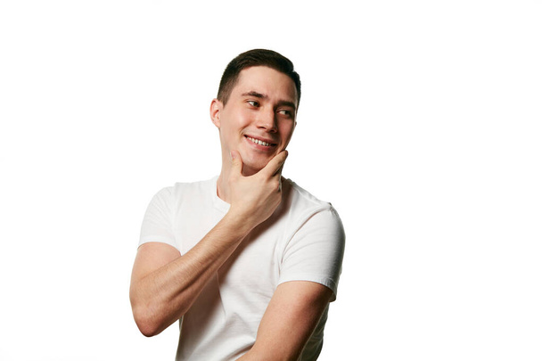 Attractive man, well-groomed guy in T-shirt smiling and touching chin looking away against white background. Concept of beauty treatment and hygiene, self-care, anti-aging procedures. Copy space. Ad - Photo, Image