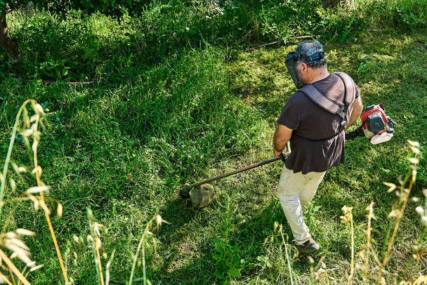 Jardineiro cortando ervas daninhas no jardim com cortador de grama corda. Homem usando cortador de grama no quintal, cortando grama verde. - Foto, Imagem