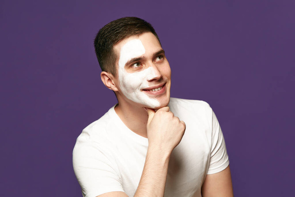 Portret wesołego mężczyzny odwracając wzrok produktem kosmetycznym, biała maska na twarzy na fioletowym tle. Koncepcja zabiegów kosmetycznych i higienicznych, pielęgnacji ciała, zabiegów anti-aging. - Zdjęcie, obraz