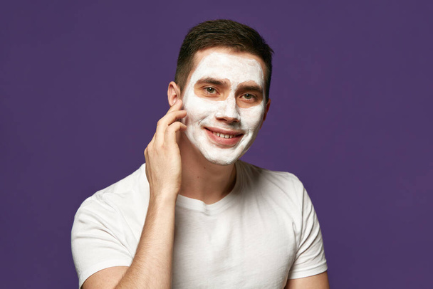 落ち着いたブルネットマン,フェイシャルケアマスク付きの魅力的な男の肖像画,紫色の背景に対する化粧品. 美容,衛生,ボディケア,アンチエイジング手順の概念. - 写真・画像