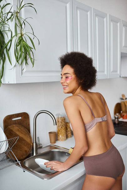 Mutlu ve kıvırcık Afro-Amerikan kadın iç çamaşırlı, gözlerinin altında pembe yamalar olan bulaşıkları yıkayan. - Fotoğraf, Görsel
