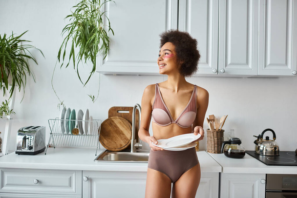 happy and curly african american woman in lingerie with pink patches under eyes holding clean dishes - Photo, Image