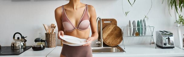 banner recortado de mulher afro-americana em lingerie segurando pratos limpos na cozinha moderna - Foto, Imagem