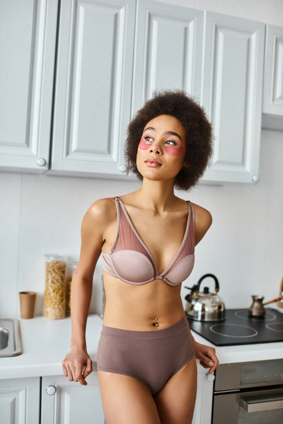 мрійлива афроамериканка в бюстгальтері з рожевими плямами під очима, стоячи на сучасній кухні - Фото, зображення