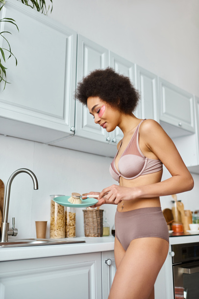 pretty african american woman in bra with pink patches under eyes washing plate with dish brush - Photo, Image