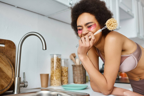 joyeuse femme afro-américaine en soutien-gorge avec des taches roses sous les yeux tenant brosse à vaisselle dans la cuisine - Photo, image