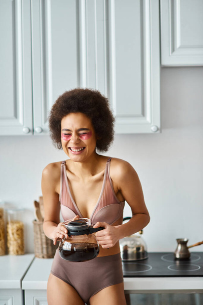 Radiante jovem afro-americana com manchas oculares segurando panela de café em uma cozinha moderna iluminada pelo sol - Foto, Imagem