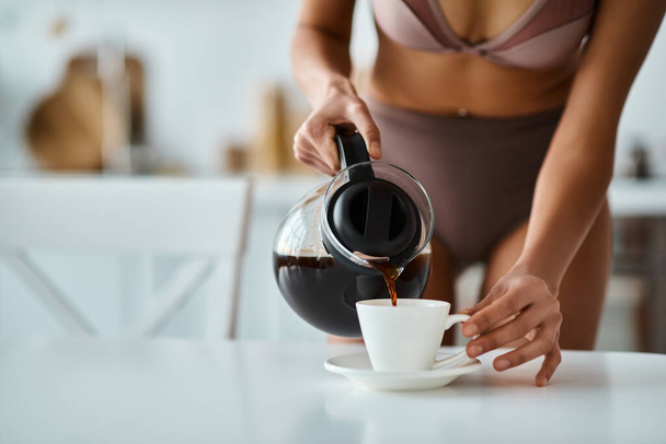 обрізаний вид афроамериканської жінки в нижній білизні, що заливає каву в порцелянову чашку на кухні - Фото, зображення