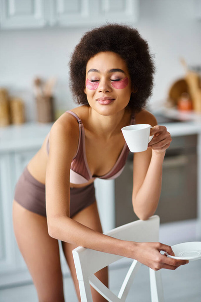 radosna Afroamerykanka z przepaskami na oczach stojąca w bieliźnie i delektująca się poranną kawą - Zdjęcie, obraz