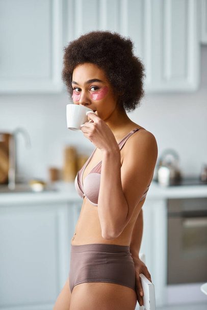 Χαρούμενη Αφροαμερικανή με μπαλώματα στα μάτια που στέκεται με εσώρουχα και πίνει πρωινό καφέ. - Φωτογραφία, εικόνα